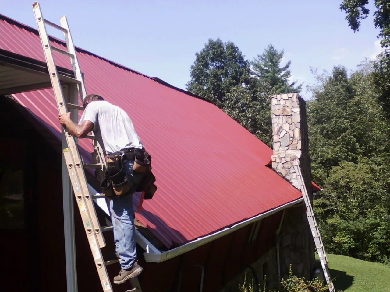 Roofer on a ladder beside a red metal roof and stone chimney — Soffit & Fascia Repair by Roofing Brothers in Badger